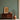 Wooden cabinet against ornate green wallpaper holds framed art, clay pot, and bowl; large vase with dried branches stands nearby.