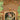 Elegant wallpaper mural shows Krishna and Radha in ornate dress beneath a peacock arch, with greenery, florals, and a monkey above.